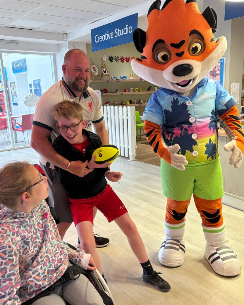 Craig, Caiden and Ella are pictured on their Haven holiday with a Tiger mascot. Ella is sitting in her wheelchair looking at the mascot, and Craig looks to be play-fighting with Caiden over a rugby ball.