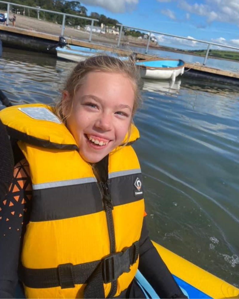 Ella is on the water in a a boat, wearing a life-vest. She is smiling.