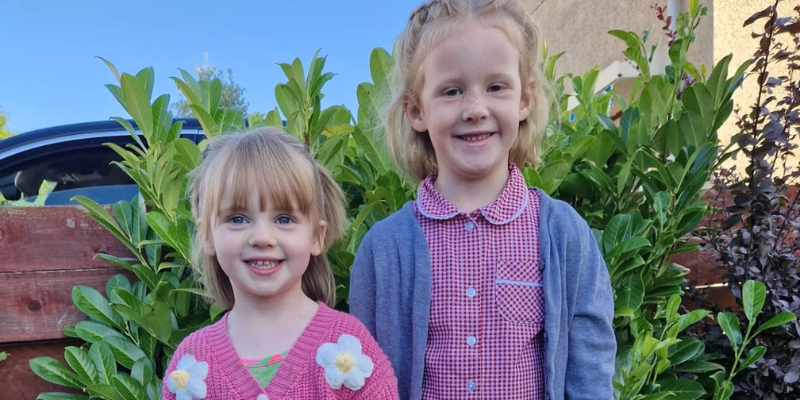 Eve and Olivia are stood in front of greenery and they are both smiling. Both girls have blond hair, smile at the camera, and are dressed with summer clothes. 