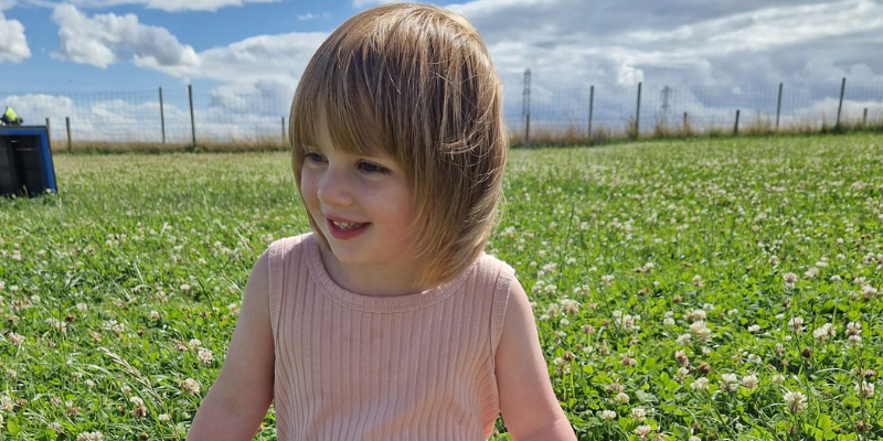 Eve is sat in a field. Her light hair and fringe is slightly blowing in the breeze. She wears a pink vest and is looking to her right, smiling.