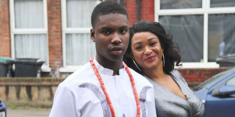 Kelly is wearing white with orange beads. His mum, Isoken, is wearing. silver, holding onto his arm and smiling, leaning towards him.