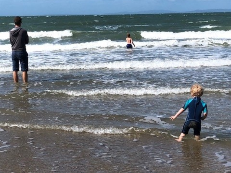 A dad in shorts stands in the shallow sea water watching his two sons in the water