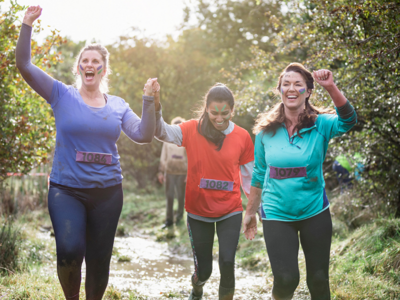 Three women are running in mud holding hands and smiling.