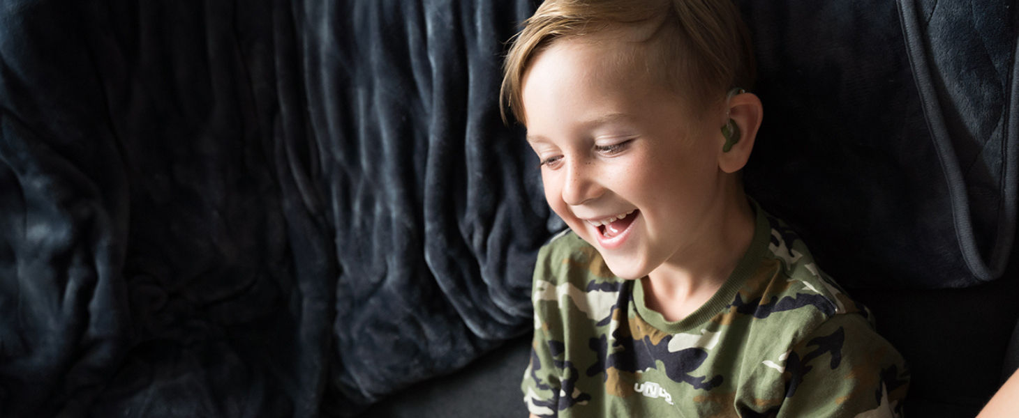 A young boy with a hearing aid, wearing a green t-shirt is sitting on a dark sofa laughing