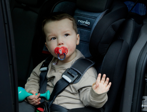 A little boy with a red dummy in his mouth is sat in a car seat waving