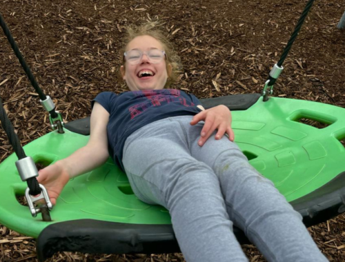 Ella is holding onto a large flat swing. She is lying on the swing and smiling widely.