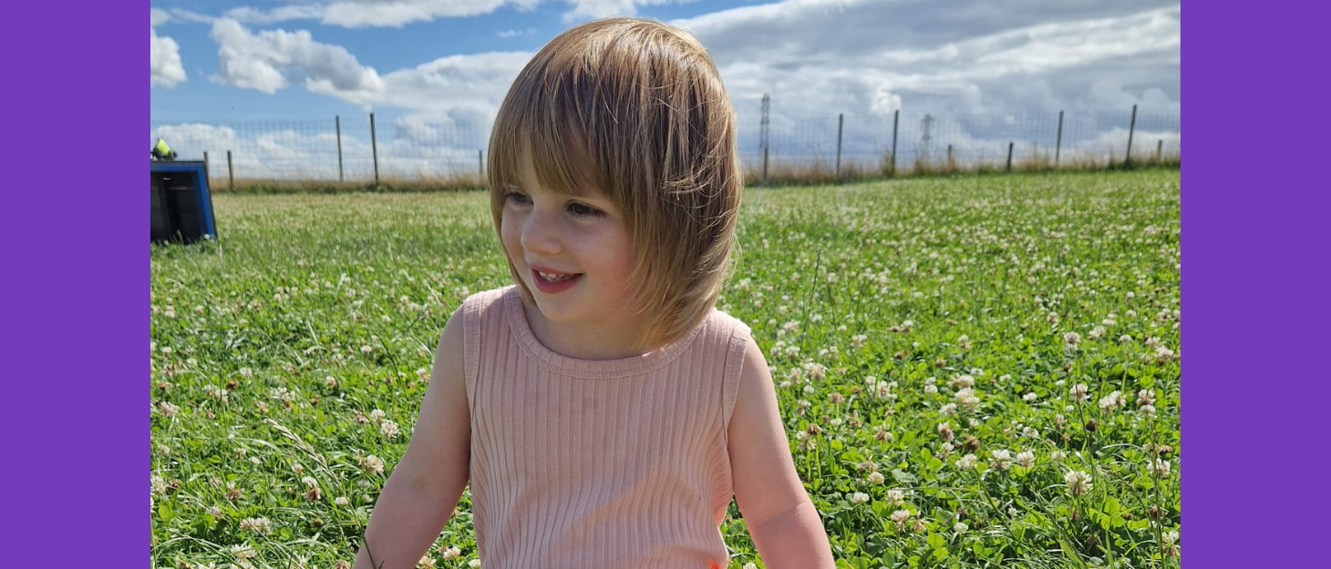 Eve is sat in a field. Her light hair and fringe is slightly blowing in the breeze. She wears a pink vest and is looking to her right, smiling.