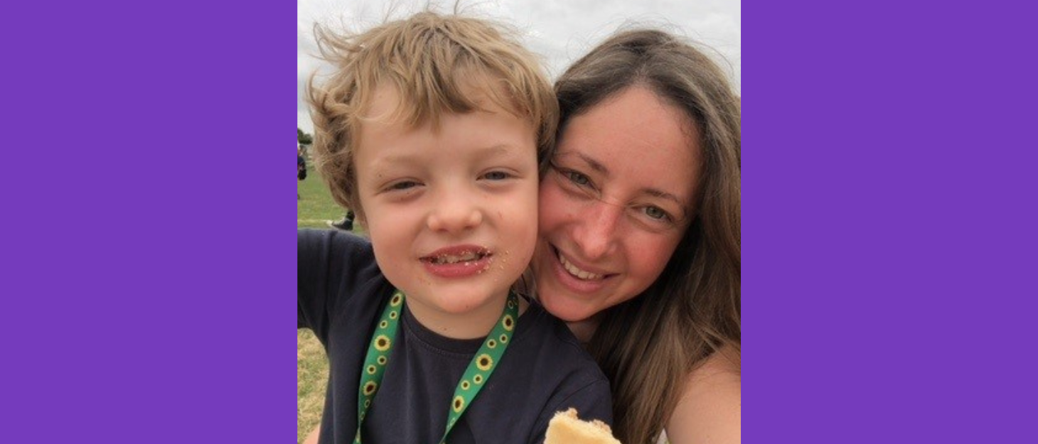 A young boy holding an ice cream cone, wearing a green lanyard smiles with his mum next to his shoulder. She has long brown hair, smiling.