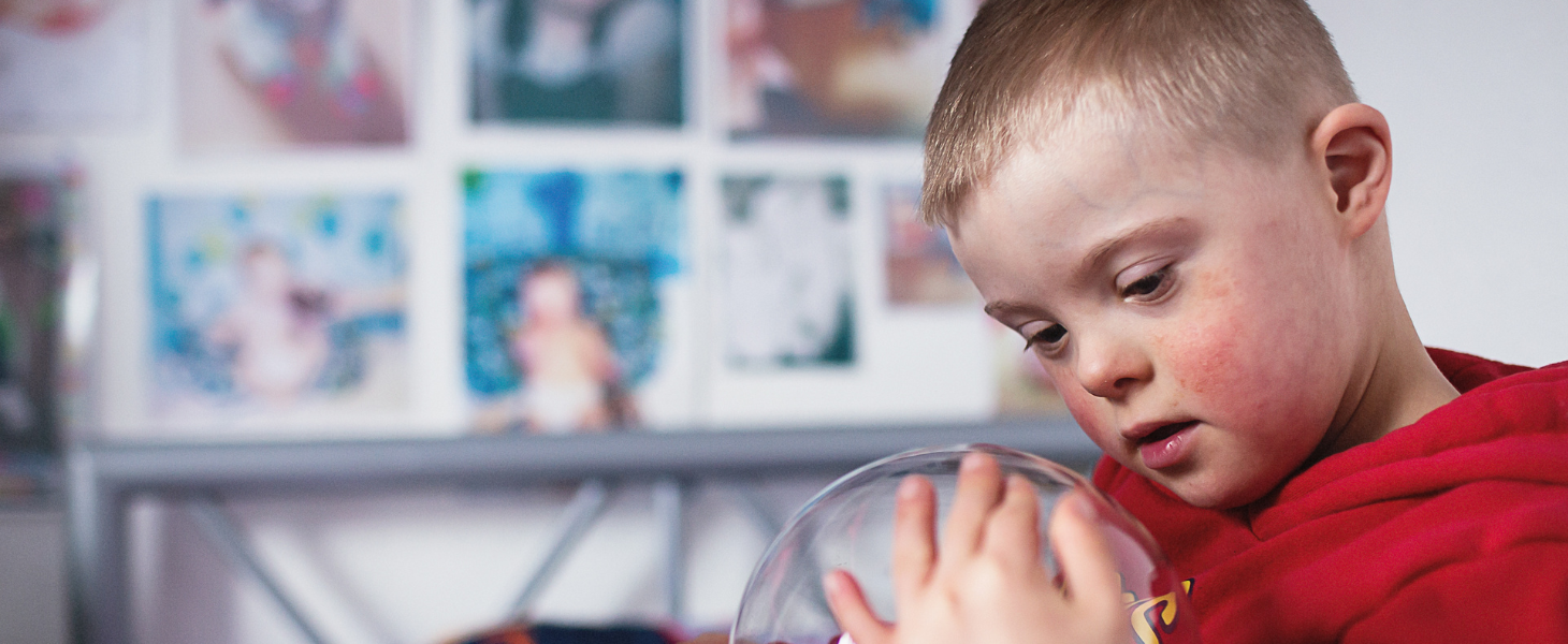 A little boy sitting on a metal frame bed, wearing a bright red hoodie is holding a plasma ball sensory toy.