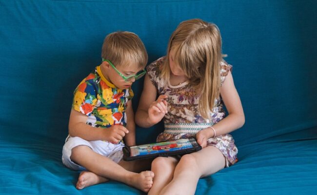 A young boy wearing green glasses sits on a blue sofa next to his sister. They're looking down at an iPad together.