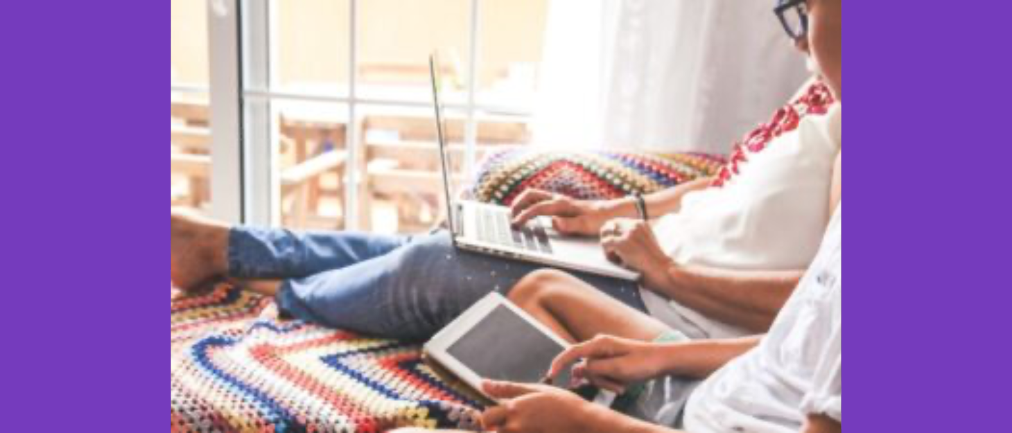 A mum sitting on a sofa with her feet up using a laptop, next to her a boy on a tablet